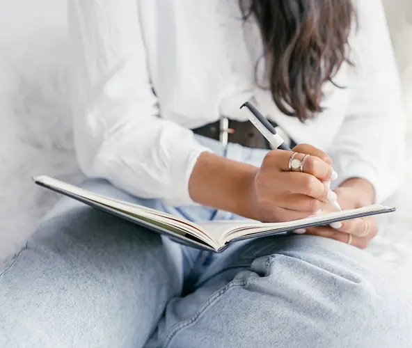 woman with jeans writing in a notebook on her lap